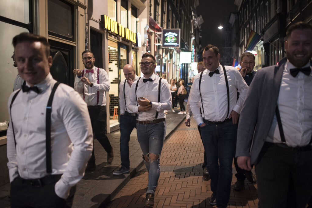 A group of guys dressed in slacks and bow ties walk down a street during an Amsterdam bachelor party. 